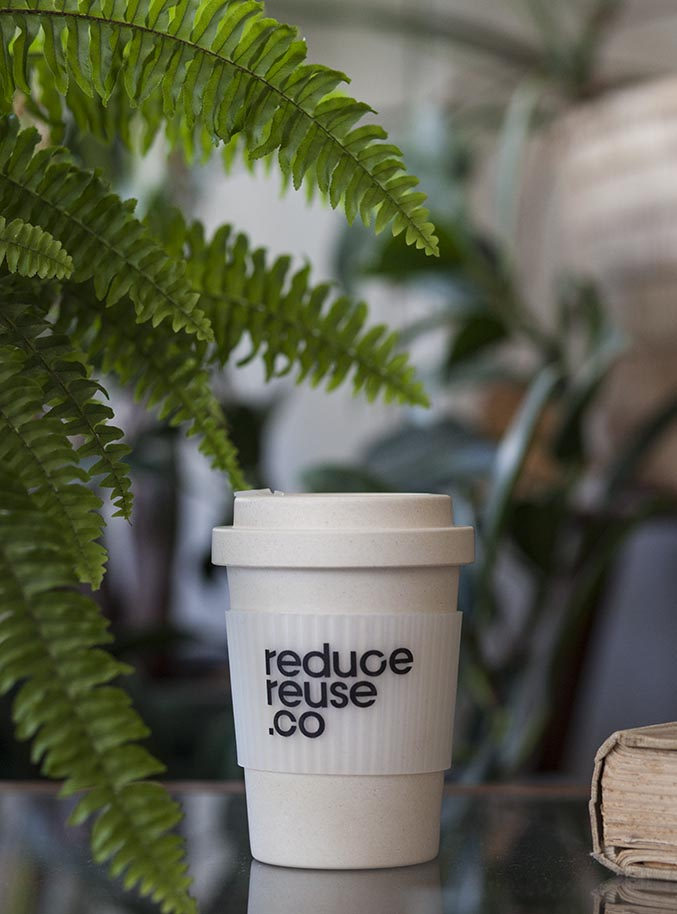 A reduce reuse .co reusable coffee cup on a glass table, framed by ferns and houseplants.